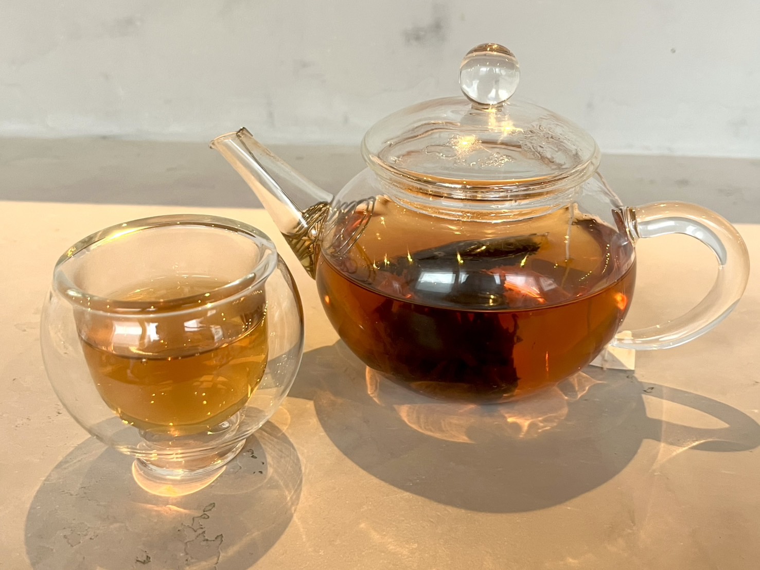ガラスのティーポットとカップに琥珀色の茶。白いカウンター上の横写真。